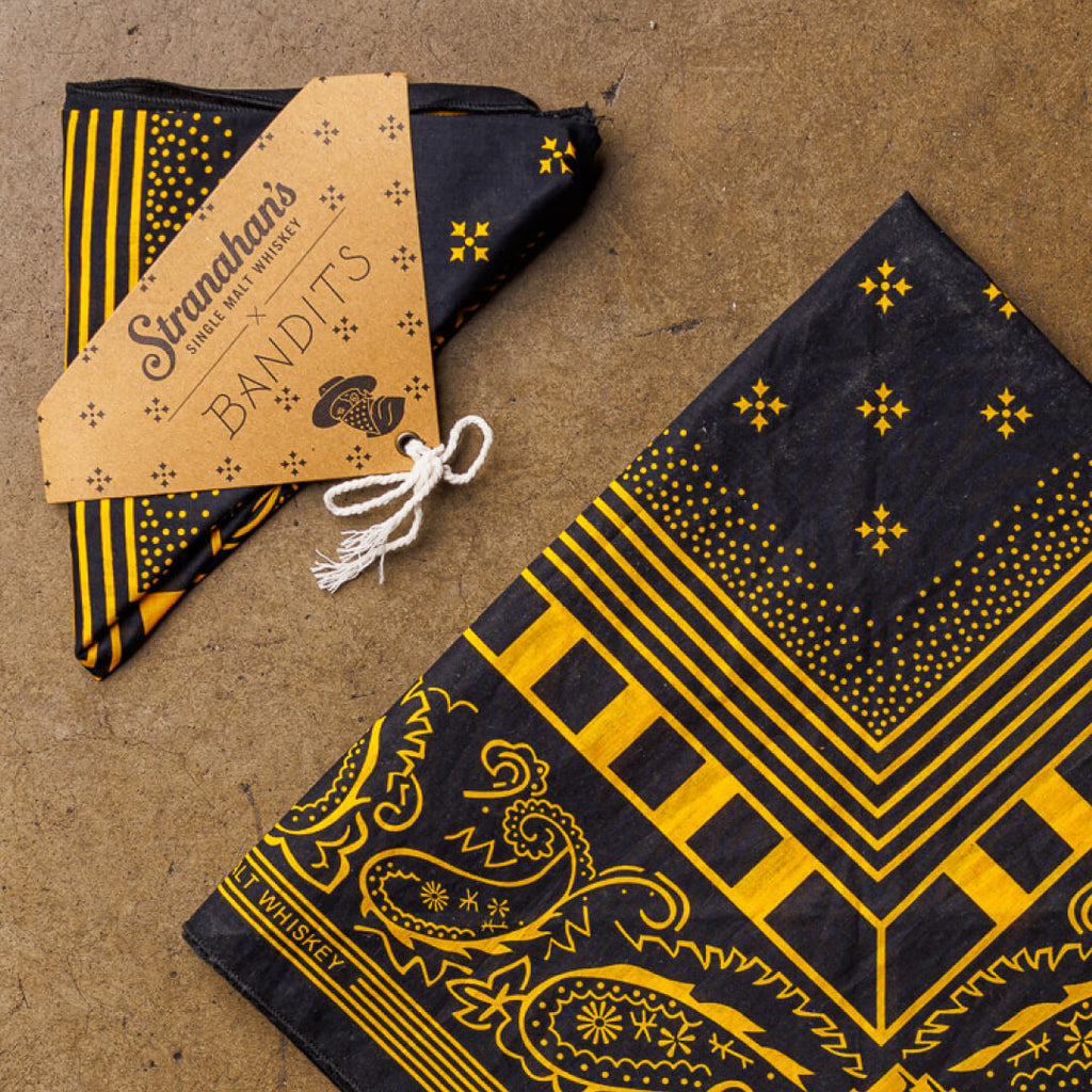 Closeup of Stranahan's Bandana by Bandits flatlay on cement surface. Yellow and black bandana is folded in a square with Bandits packaging beside it.