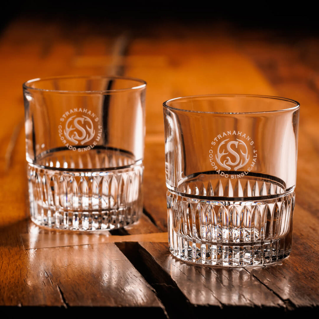 A pair of Stranahan’s Crystal Tumblers with an etched Stranahan’s logo, placed side by side on a wooden surface, reflecting light for a sleek and elegant look.