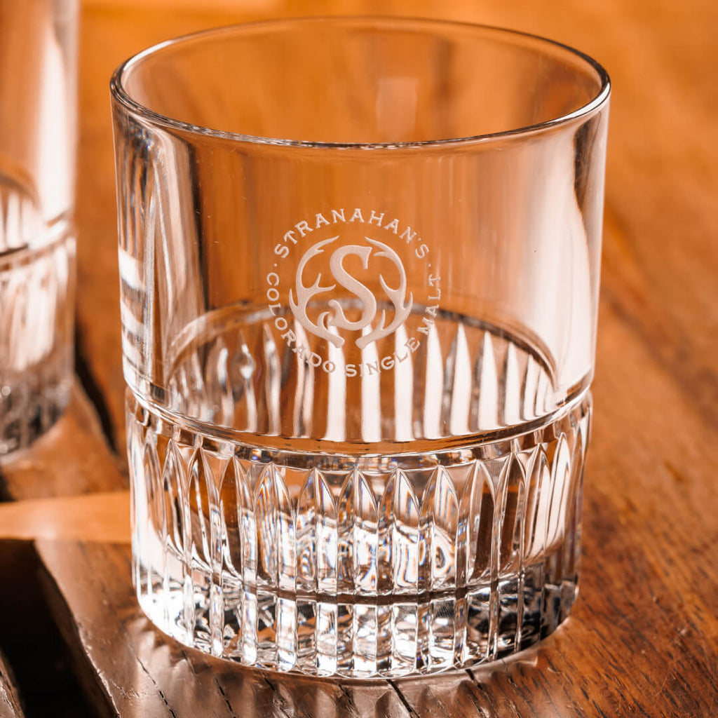 A pair of Stranahan’s Crystal Tumblers with an etched Stranahan’s logo, placed side by side on a wooden surface, reflecting light for a sleek and elegant look.