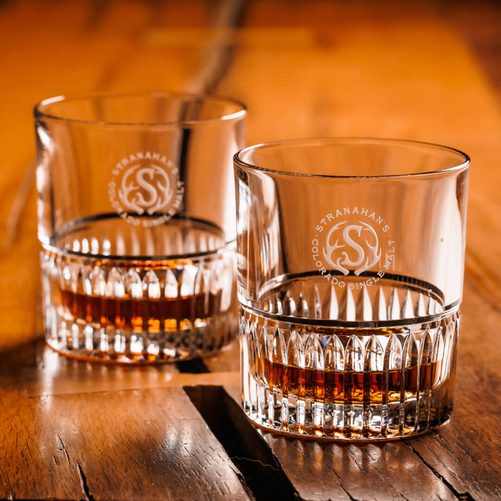 A pair of Stranahan’s Crystal Tumblers with an etched Stranahan’s logo, placed side by side on a wooden surface, reflecting light for a sleek and elegant look.