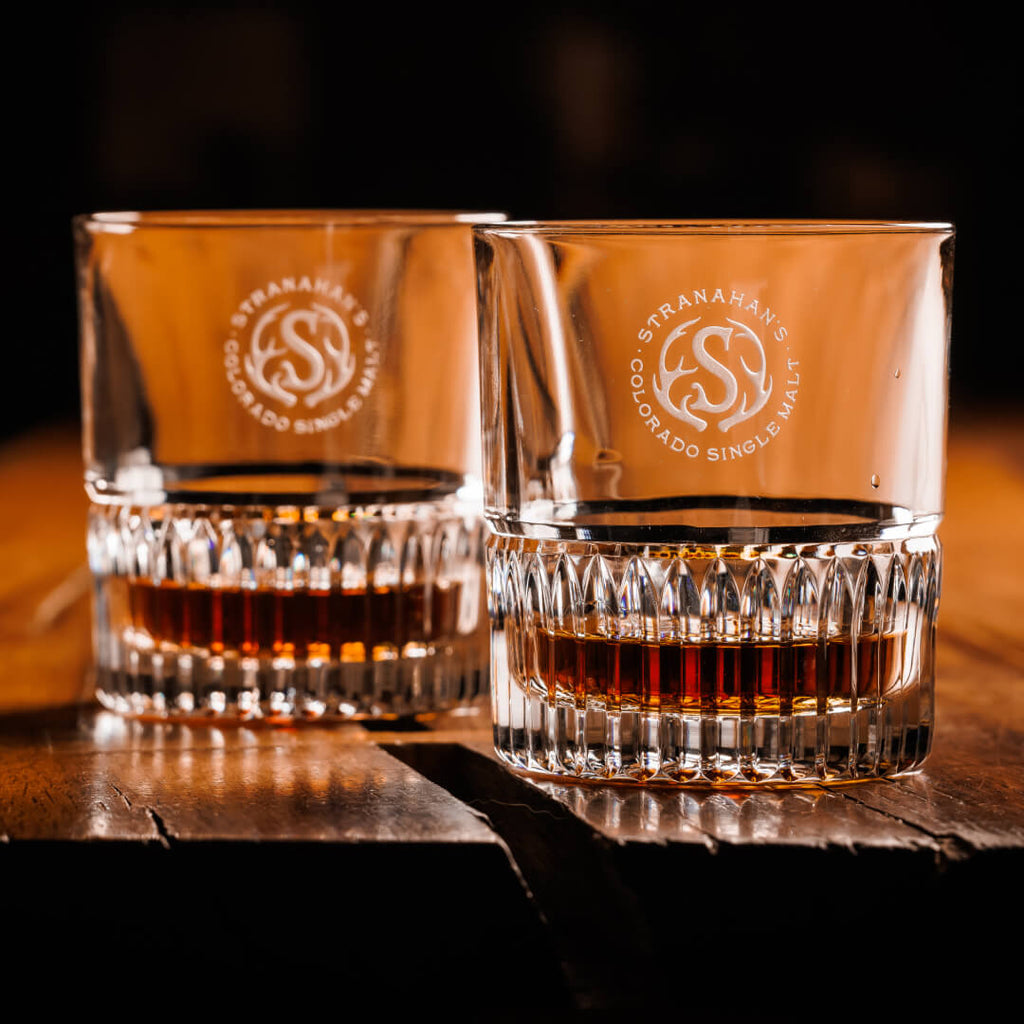 A pair of Stranahan’s Crystal Tumblers with an etched Stranahan’s logo, placed side by side on a wooden surface, reflecting light for a sleek and elegant look.