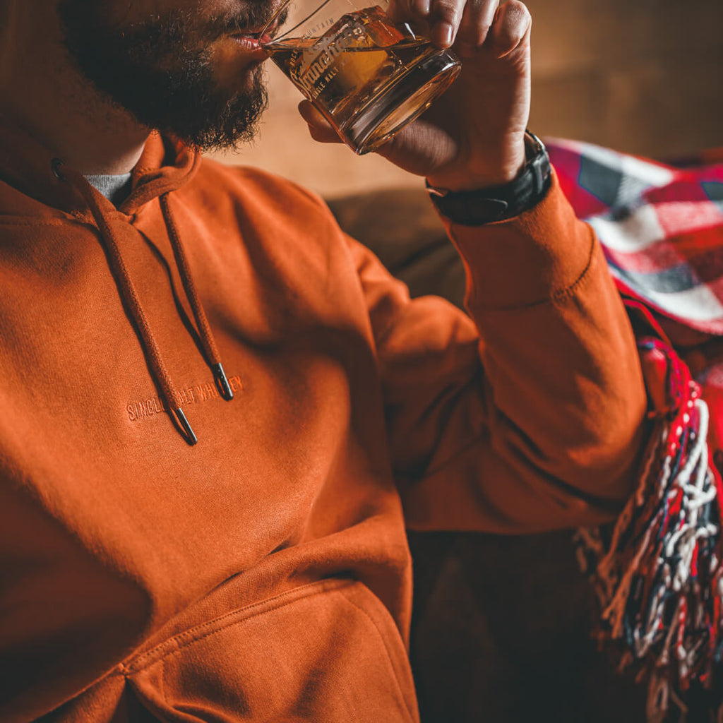 Closeup shot of man wearing a burnt orange Stranahan's Embroidered Clay Sweatshirt while drinking a whiskey cocktail