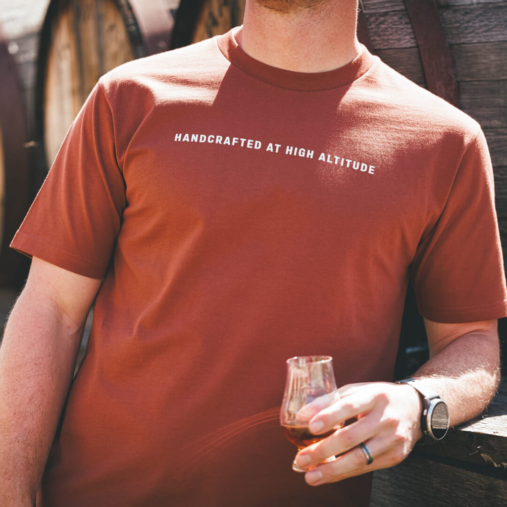 Man wearing rustic clay colored Stranahan's Handcrafted at High Altitude Tee while holding dram of Stranahan's Whiskey