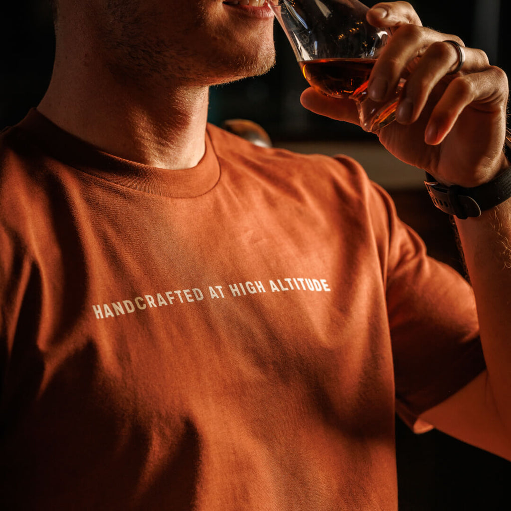Man wearing rustic clay colored Stranahan's Handcrafted at High Altitude Tee while drinking dram of Stranahan's Whiskey