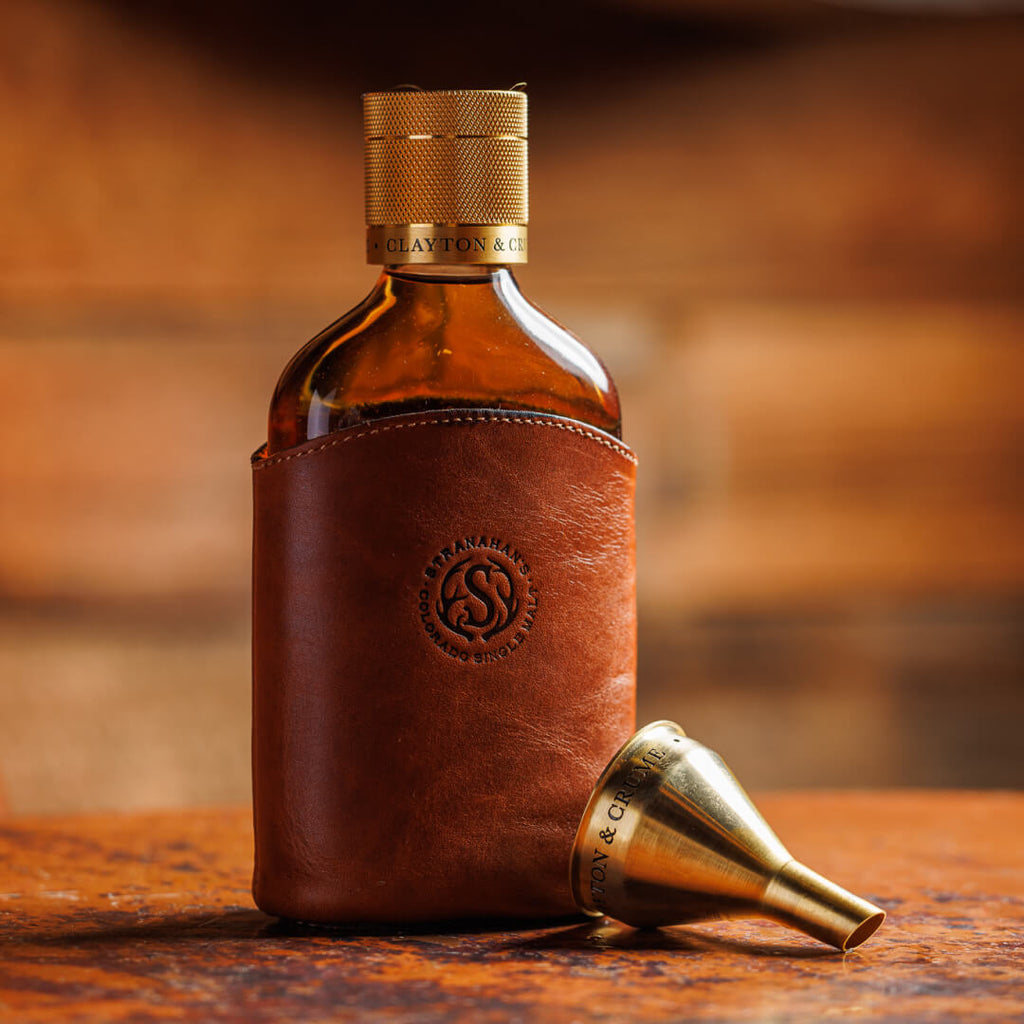 Leather Wrapped Glass Flask stamped with the Stranahan's logo with gold screw top and gold funnel for pouring placed next to it