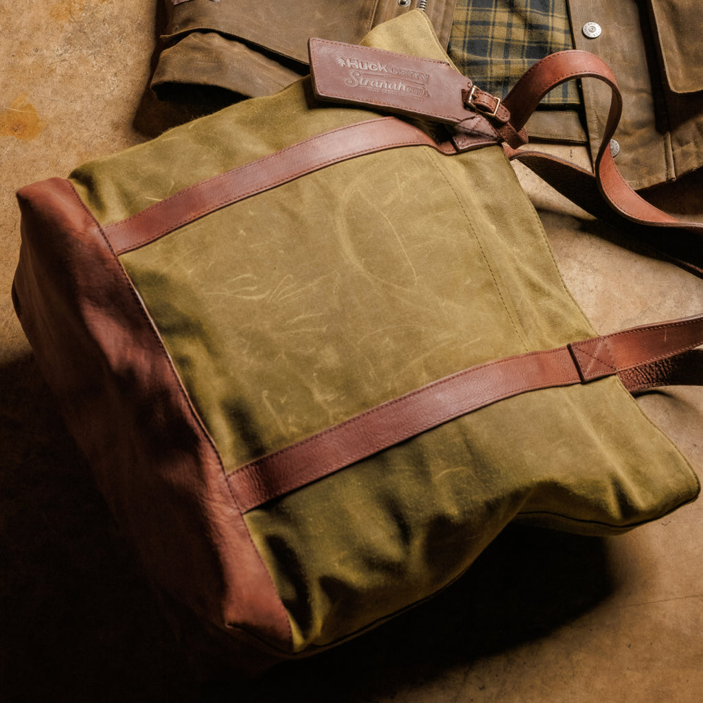 Person holding a green canvas and leather tote bag with a wooden barrel in the background.