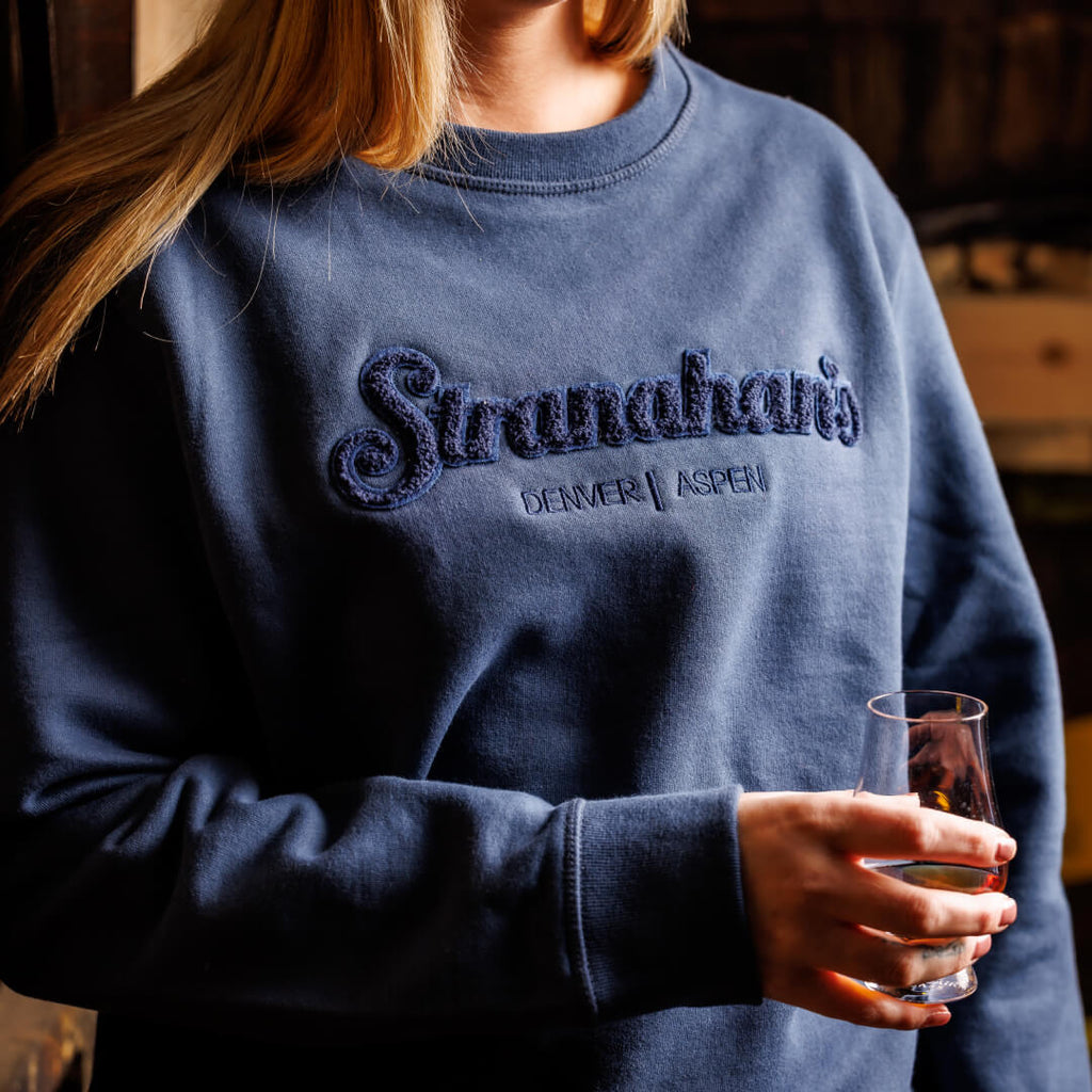 Three-quarter view of woman wearing navy sweatshirt with a chenille Stranahan's logo patch embroidered with Denver and Aspen locations
