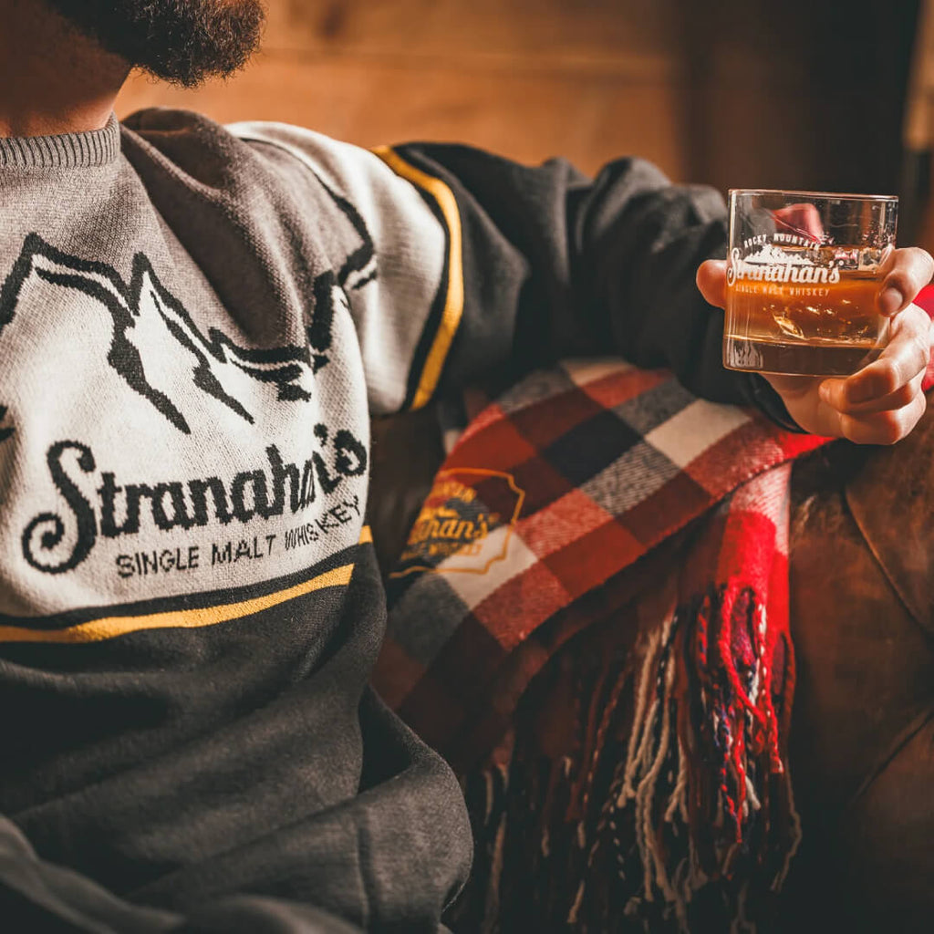 Man wearing Stranahan's Shinesty Mountain sweater while sitting on leather couch and holding a whiskey cocktail