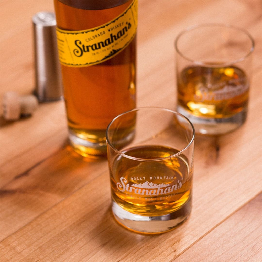 Bottle of Stranahan's Colorado Whiskey with two glasses filled with whiskey, showcasing the Rocky Mountain Stranahan's logo, on a wooden surface.