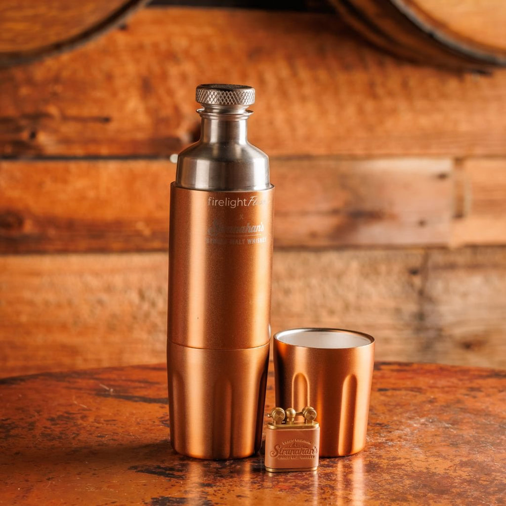 Stranahan's copper flask, cup, and leather-bound-lighter placed on a wooden surface with a barrel background.