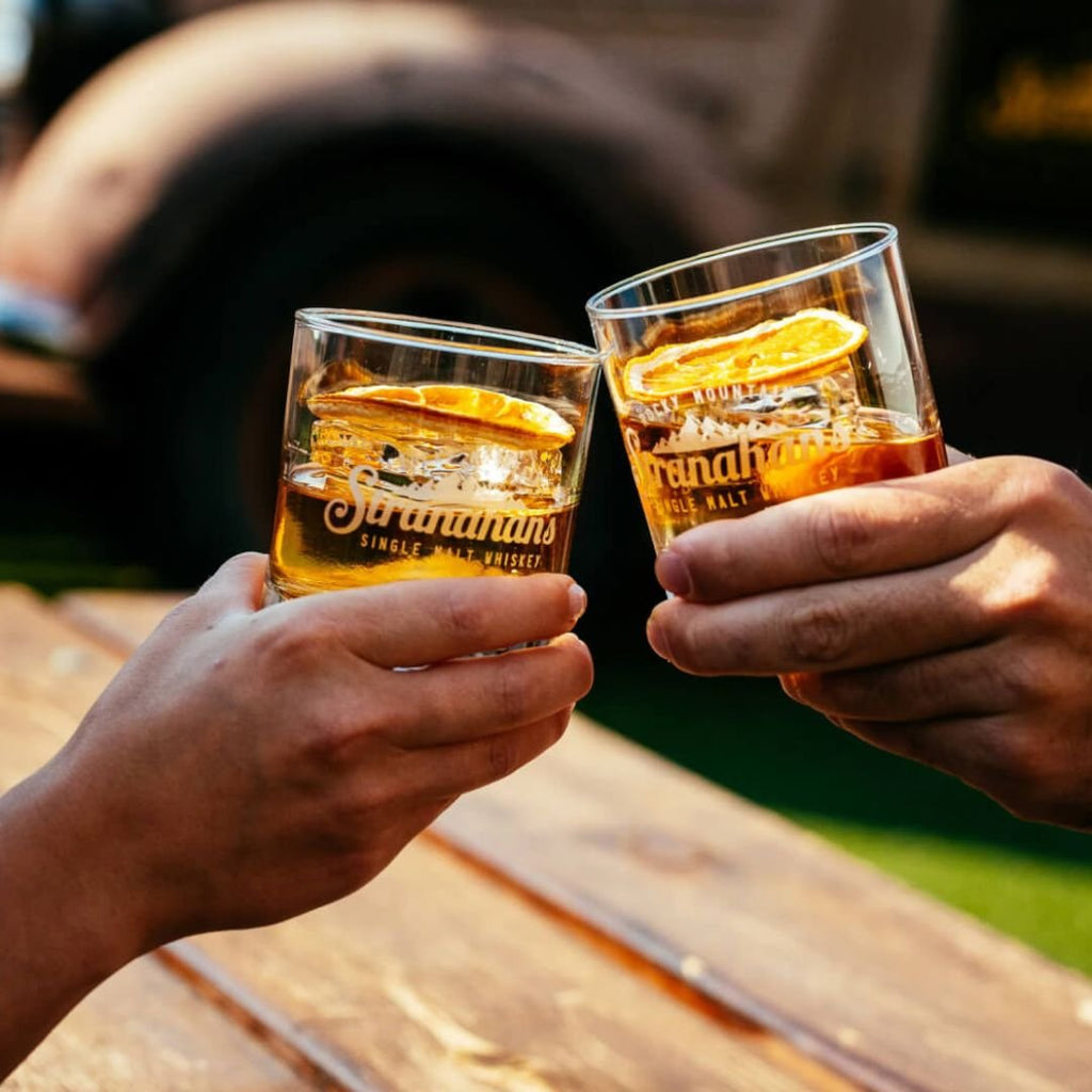 Two hands clinking whiskey glasses with orange slices, featuring Stranahan's Single Malt Whiskey logo, in a warm outdoor setting.