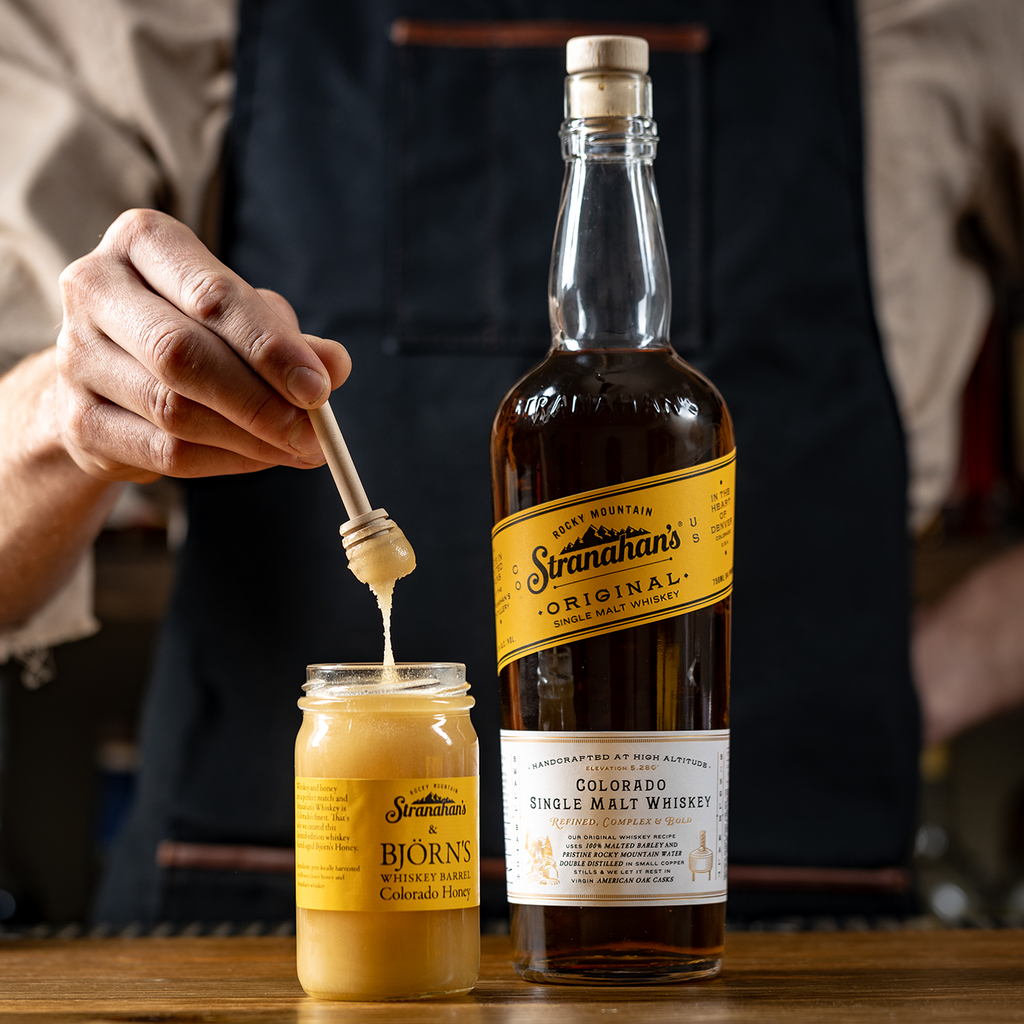 Honey wand dipped into open jar of Stranahan's whiskey barrel Colorado honey next to bottle of Stranahan's Original Whiskey.